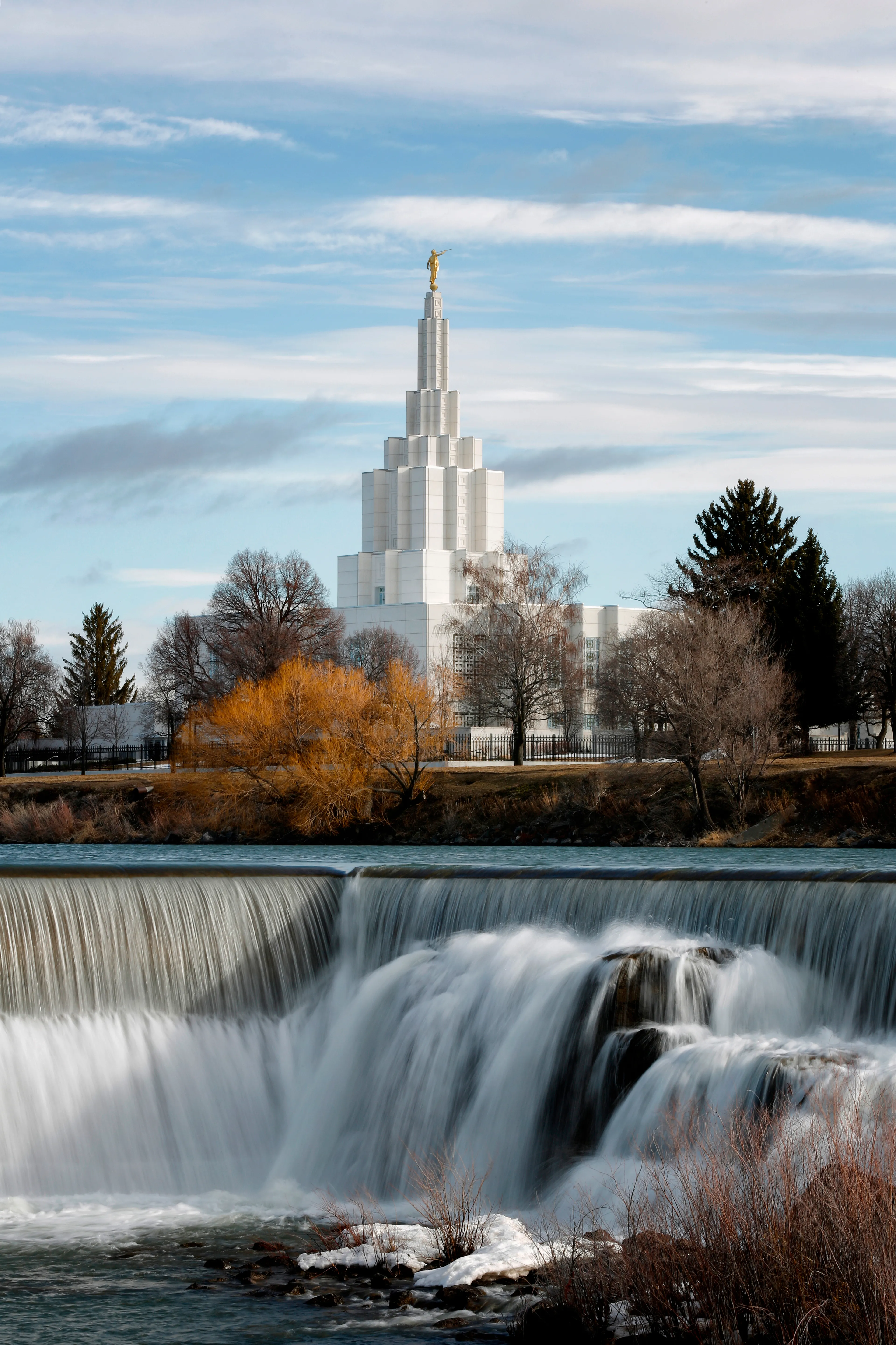 Exterior of the Idaho Falls Idaho temple with a river and waterfall in the foreground.