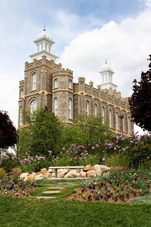 An image of the Logan Utah Temple taken from the bottom of the hill on a sunny day, with flowers around the temple and a blue sky overhead.