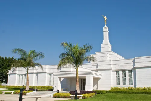 The front of the Oaxaca Mexico Temple on a sunny day, with palm trees on either side of the pathway leading up to the temple.