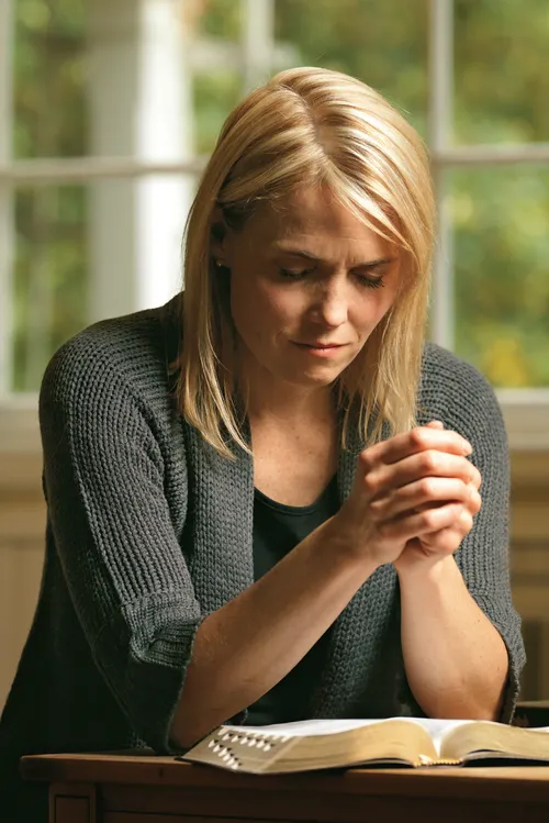 A woman sitting at a desk with the scriptures open in front of her.  She has her hands clasped and eyes closed in prayer.