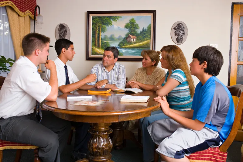 Two elder missionaries sit with a family around their kitchen table teaching them about the Restoration of the Church of Jesus Christ