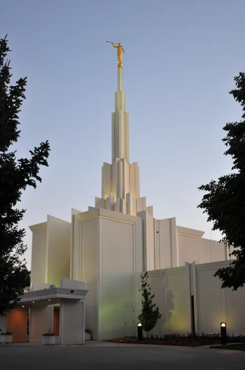 A front side view of the Denver Colorado Temple in the evening, with large trees on either side.