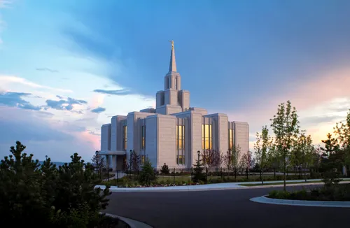 Clouds and a sunset behind the Calgary Alberta Temple, with a road and some vegetation in the foreground.