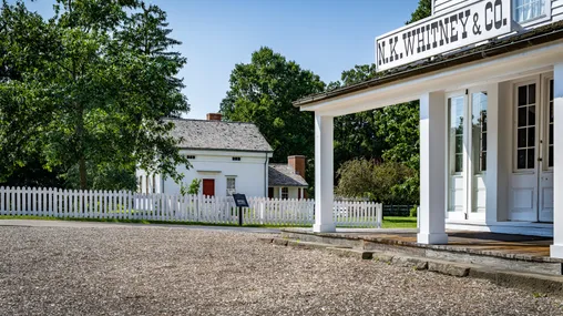 A white building with a pillared porch. A sign on the porch roof reads "N.K. Whitney & Co." A white home stands in the background.