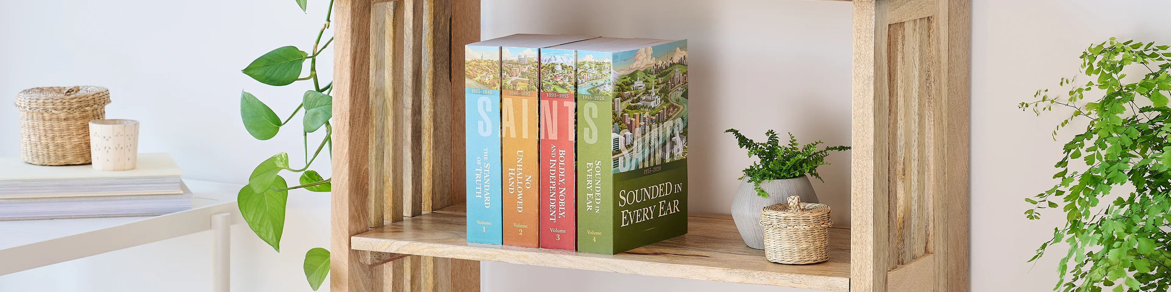 Picture of the book series, 'Saints' volume 1-4 (English edition). Four volumes are arranged on the middle shelf of a wooden bookshelf along with plants.