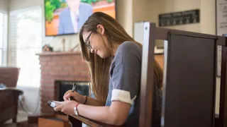 Young Vietnamese woman wearing eyeglasses, writing and studying scriptures while watching General Conference. (horiz)