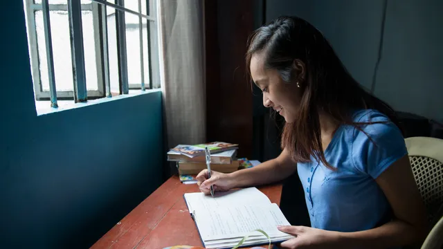 A young girl sits at a desk. She has a stack of books near her and what appears to be a journal on the desk. She is looking through it and writing. Her desk is near a window.