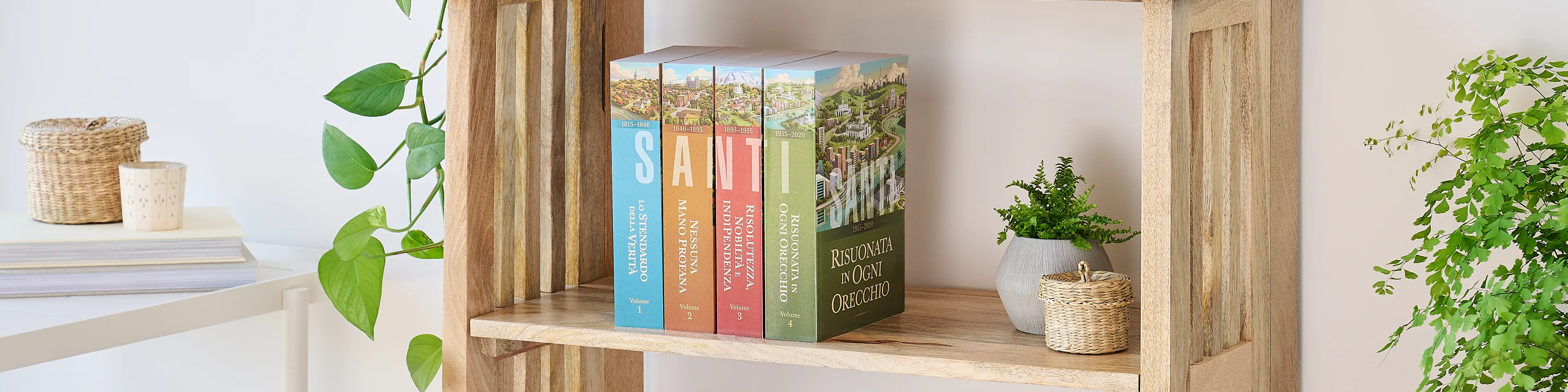 Picture of the book series, 'Saints' volume 1-4 (Italian edition). Four volumes are arranged on the middle shelf of a wooden bookshelf along with plants.