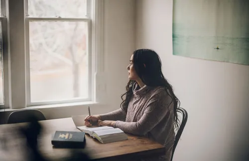 a young adult sitting at a table with her scriptures and staring out the window
