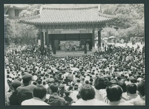 A crowd of hundreds surrounds a traditional Korean stage where young men stand with guitars, a keyboard, and two clowns. A sign behind them reads “New Horizon” and “Tender Apples.”