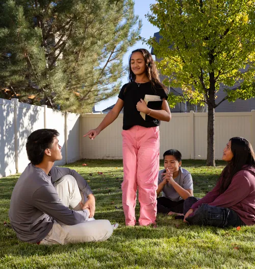 Girl standing with her eyes closed holding the scriptures, pointing at children sitting around her